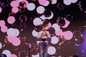 Bright stage with a speaker at KYCK 2026 event, colourful abstract background with pink and purple light effects, promoting youth leadership and community engagement.