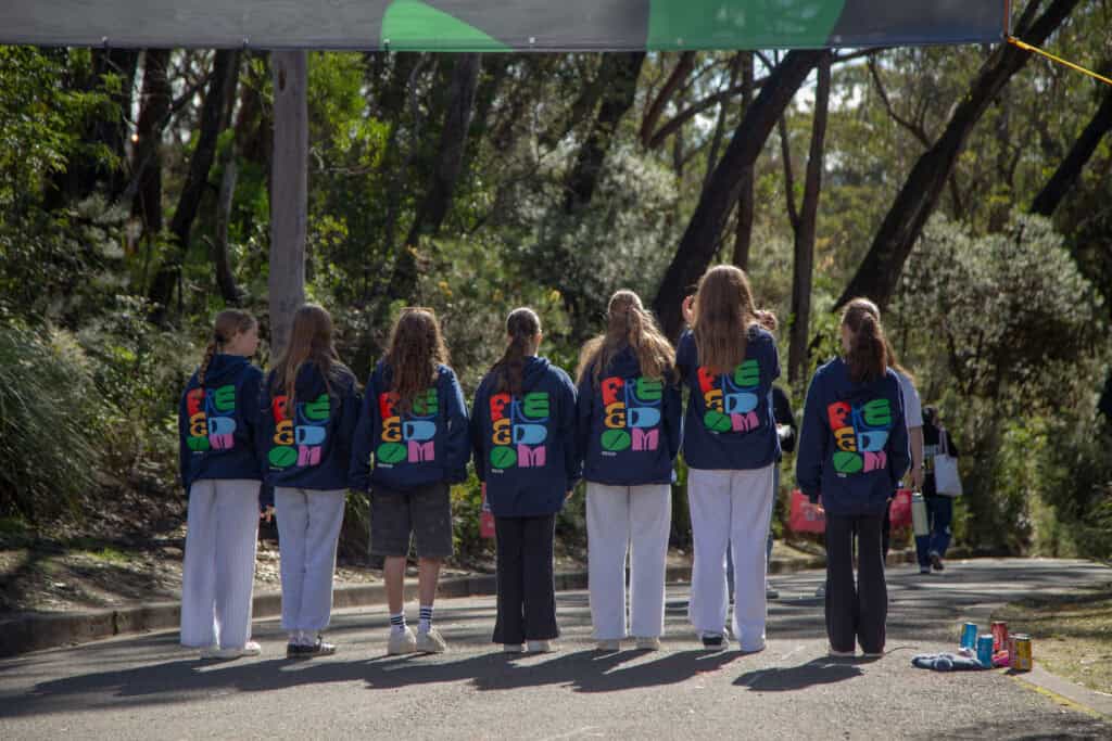 Colourful youth groups in navy hoodies with "FREEDOM" text, standing outdoors in a lush, wooded area during KYCK 2026 event in Australia, engaging in a camp activity.