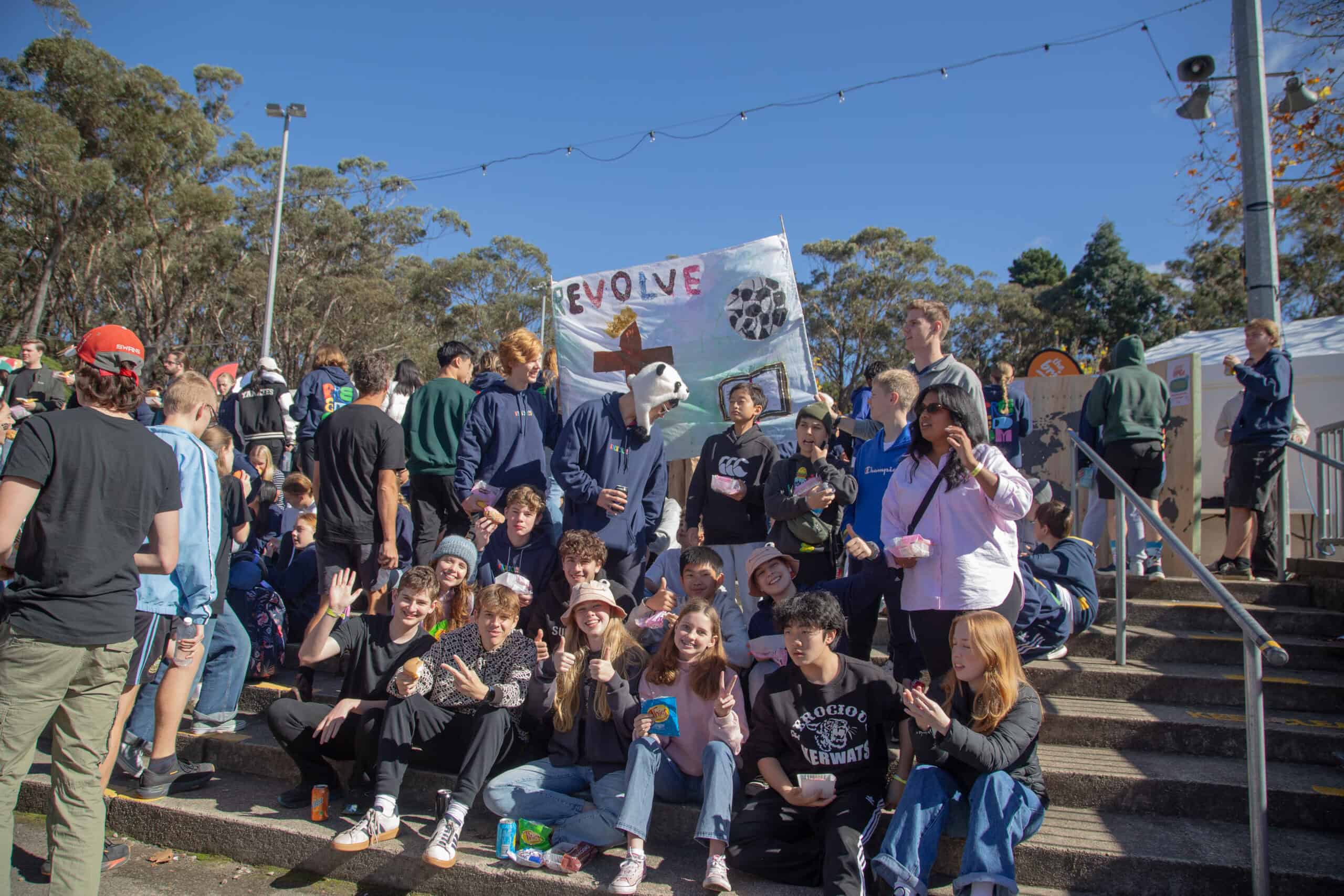 Bright outdoor scene at KYCK 2026 youth event with diverse participants, some wearing hoodies and hats, gathering on stairs, holding snacks and a colourful sign “Revolve” amidst trees and clear blue sky.