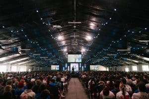 Large indoor event at KyCK 2026 with a packed audience, stage, and big screens in a spacious hall decorated with string lights, focusing on youth Christian conference.
