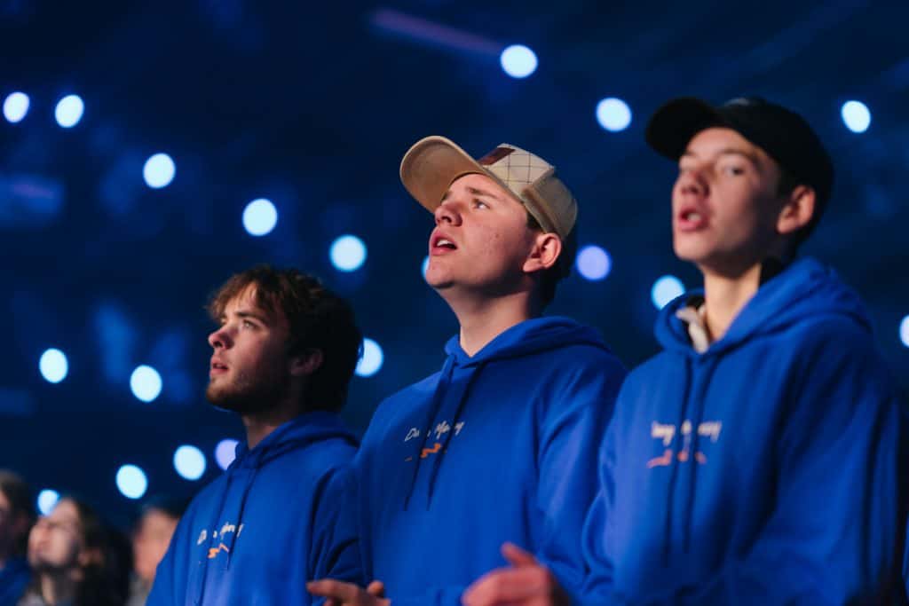 Young people attending KYCK 2026 youth conference, wearing blue hoodies, engaged in worship or prayer, with a vibrant stage background and bright lights.