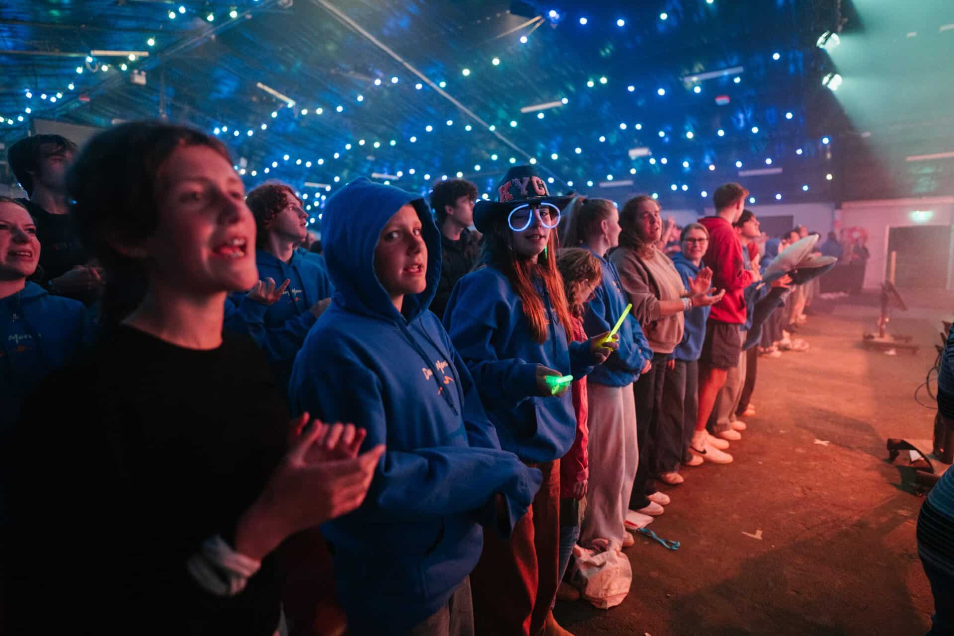Group of young people attending KYCK 2026 youth event, standing in a line with glow sticks, under vibrant stage lighting, engaging in a lively indoor gathering.