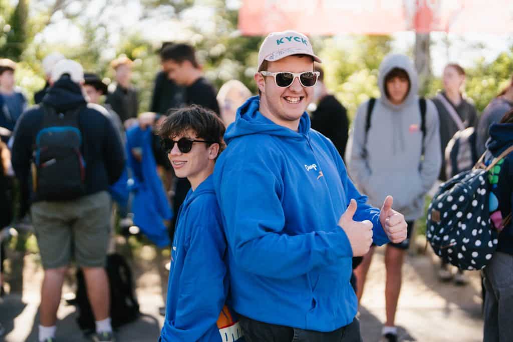 Group of excited young people at KYCK 2026 youth event, wearing blue hoodies and caps, outdoors with trees and other participants in the background.
