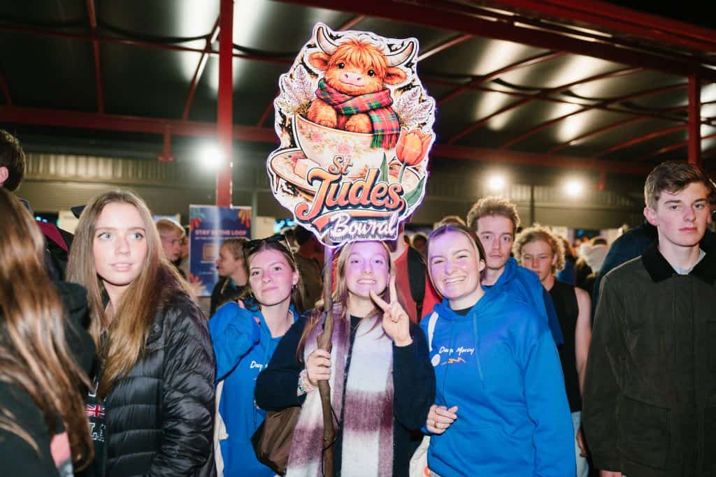 Group of young people at KYCK 2026 youth event, holding a St Jude's Bowral sign, inside a large hall with bright lighting and red structural beams.