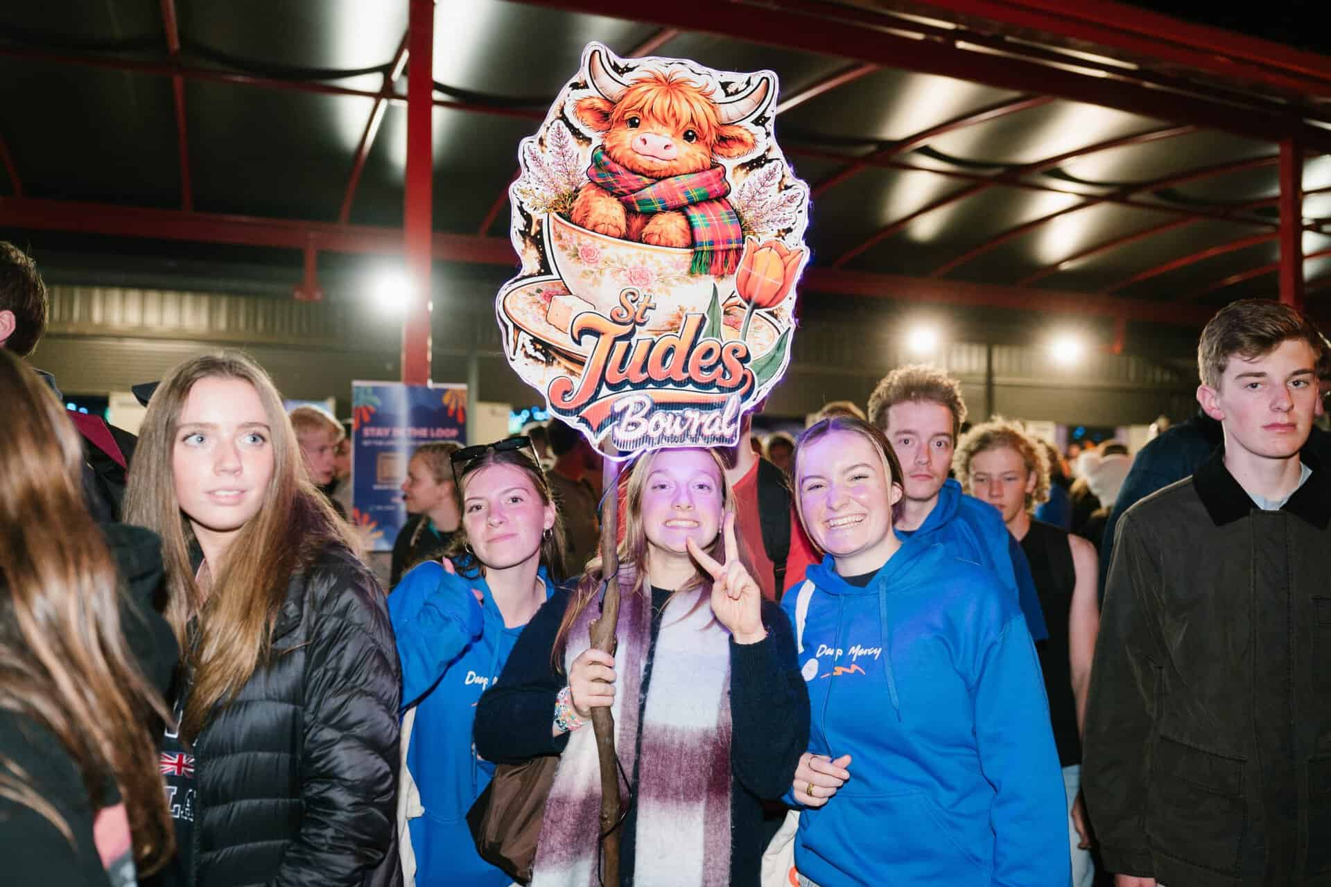 Group of young people at KYCK 2026 youth event, holding a St Jude's Bowral sign, inside a large hall with bright lighting and red structural beams.