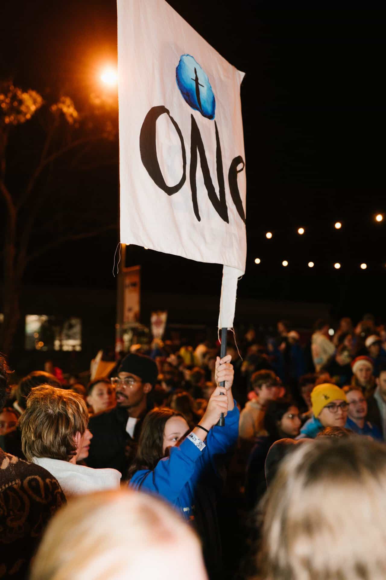 A young person holding a "One" sign at KYCK 2026 youth conference, with a large crowd and evening lights in the background.