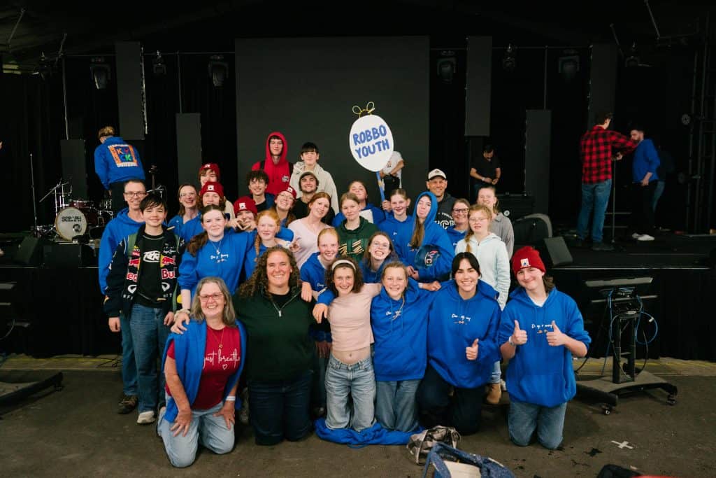 Group of young people and leaders at KYCK 2026 event, standing on stage with a "ROBO YOUTH" sign, smiling and giving thumbs up, in a large indoor venue with stage equipment.