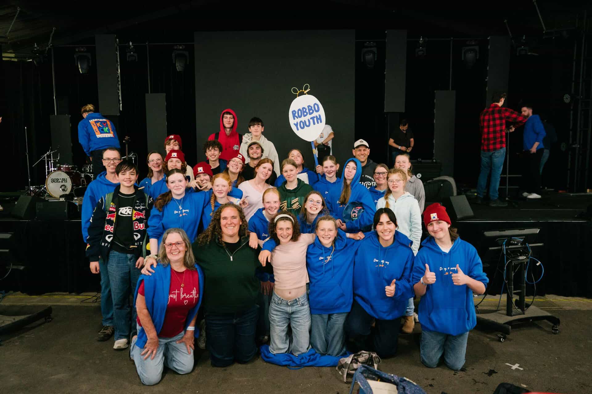 Group of young people and leaders at KYCK 2026 event, standing on stage with a "ROBO YOUTH" sign, smiling and giving thumbs up, in a large indoor venue with stage equipment.