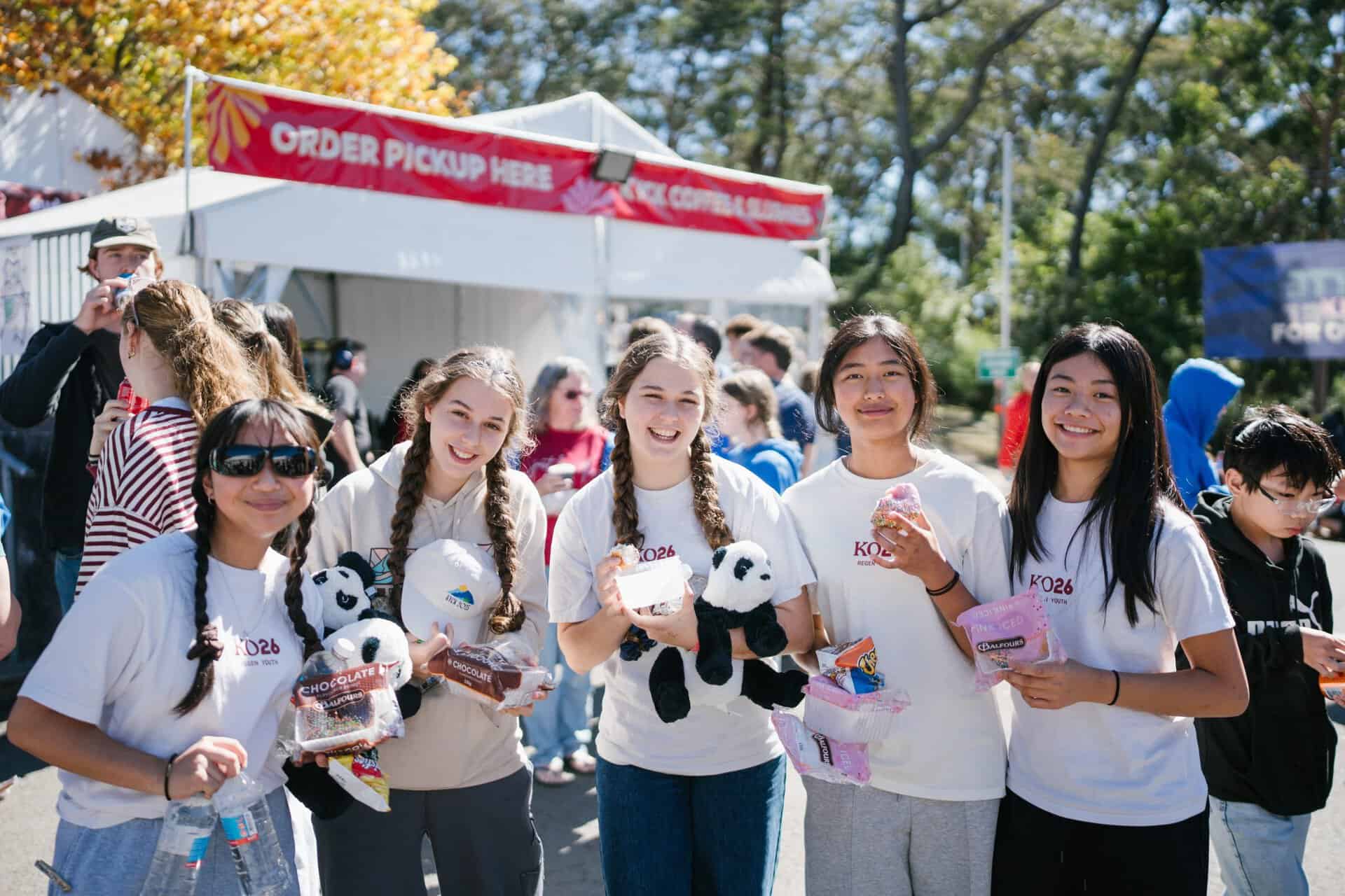 Group of smiling young people at KYCK 2026 youth event, holding panda plush toys and snacks, outdoors with a registration tent in the background.