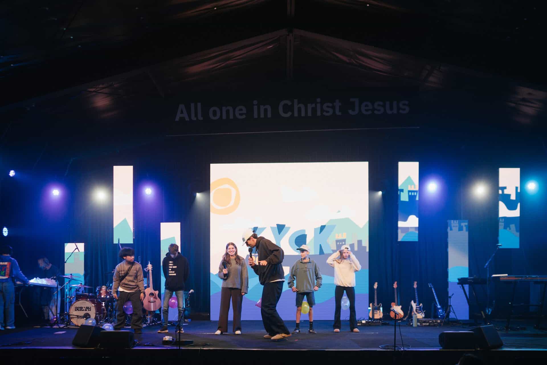 Youth conference stage at KYCK 2026 with speakers and musicians performing under a large screen displaying "All one in Christ Jesus".