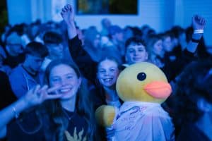 Group of excited young people at KYCK 2026 youth conference, smiling and raising hands, with a person in a duck costume, in a large indoor event hall.