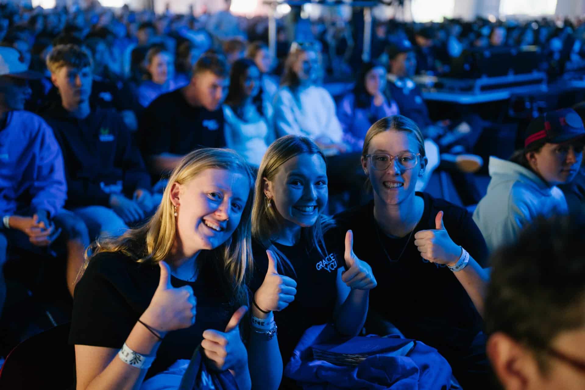 Group of excited young people at KYCK 2026 youth conference, sitting in an auditorium with blue lighting, smiling and giving thumbs up.