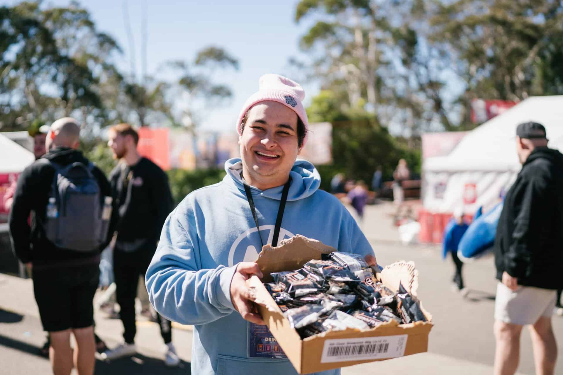 Young man smiling at KYCK 2026 youth event, holding a box of snacks outdoors with other attendees in the background.