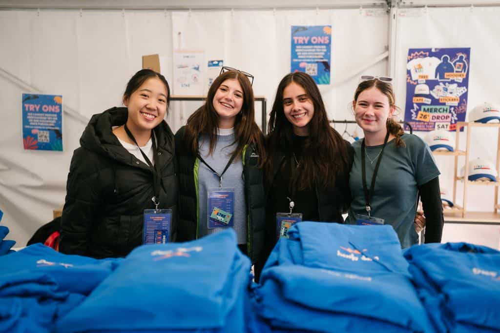 Group of four smiling young women at KYCK 2026 youth event, standing behind registration table with event merchandise and promotional materials.
