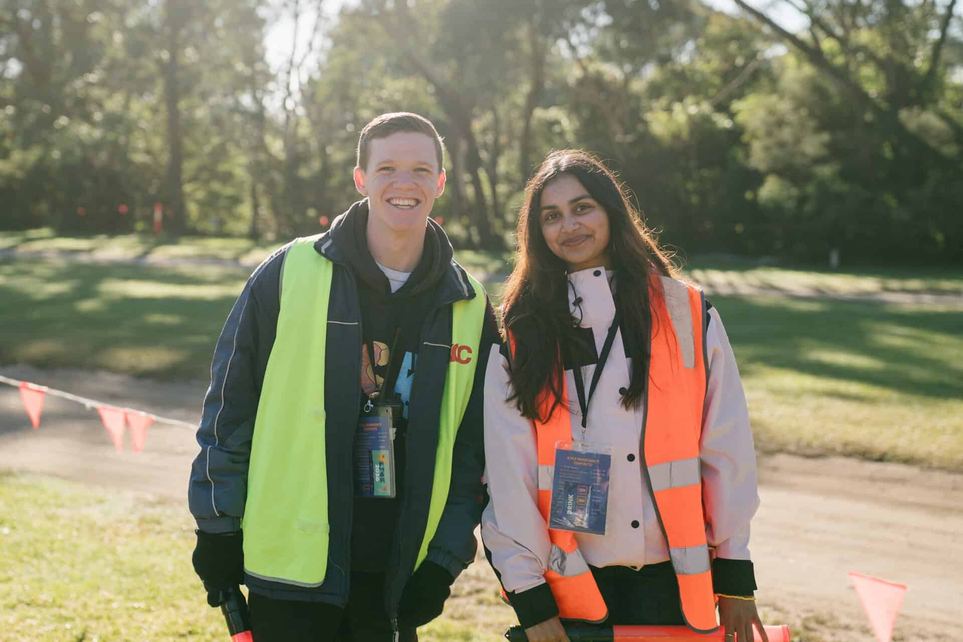 Two smiling youth volunteers at KYCK 2026 outdoor event, wearing high-visibility vests, standing in a park with trees and grass, promoting youth engagement and community activities.
