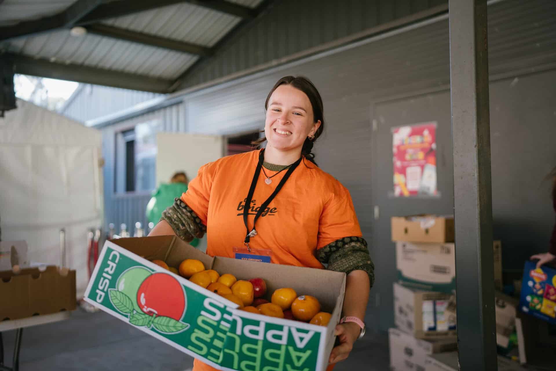 Young volunteer smiling while holding a box of fresh oranges at KYCK 2026 event, promoting community service and youth engagement.