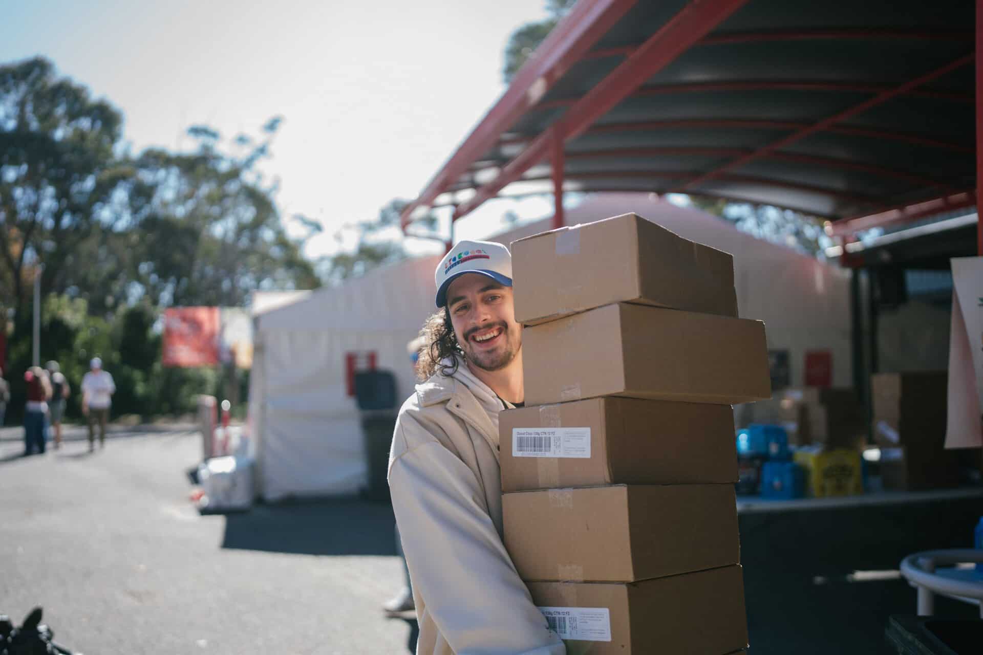 Young volunteer smiling and holding stacked boxes at KYCK 2026 event, outdoor setting with tents and people in background.