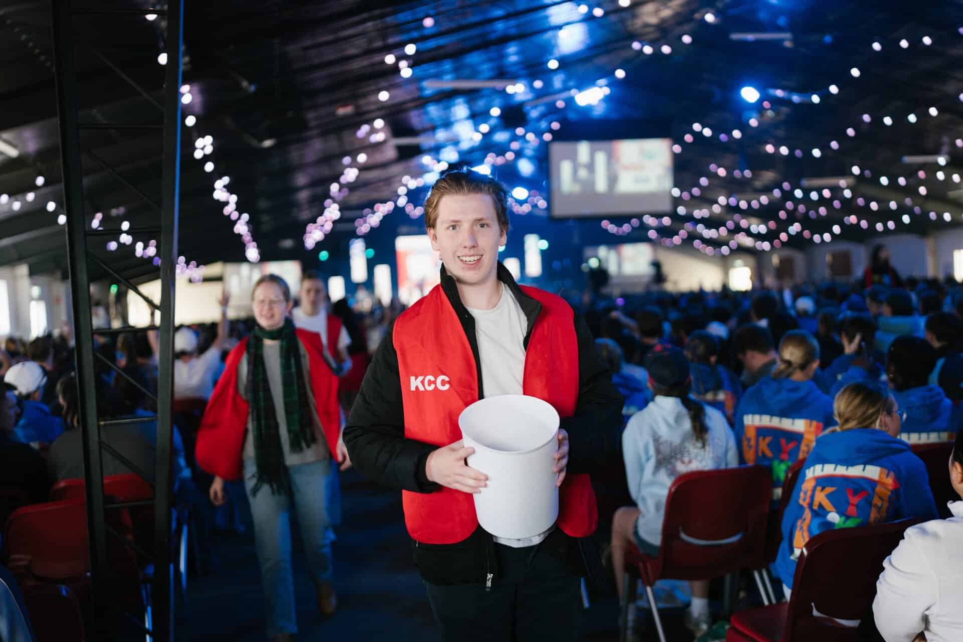 Young man smiling at KYCK 2026 youth conference, holding a donation bucket, with a lively crowd and colourful lights in the background.