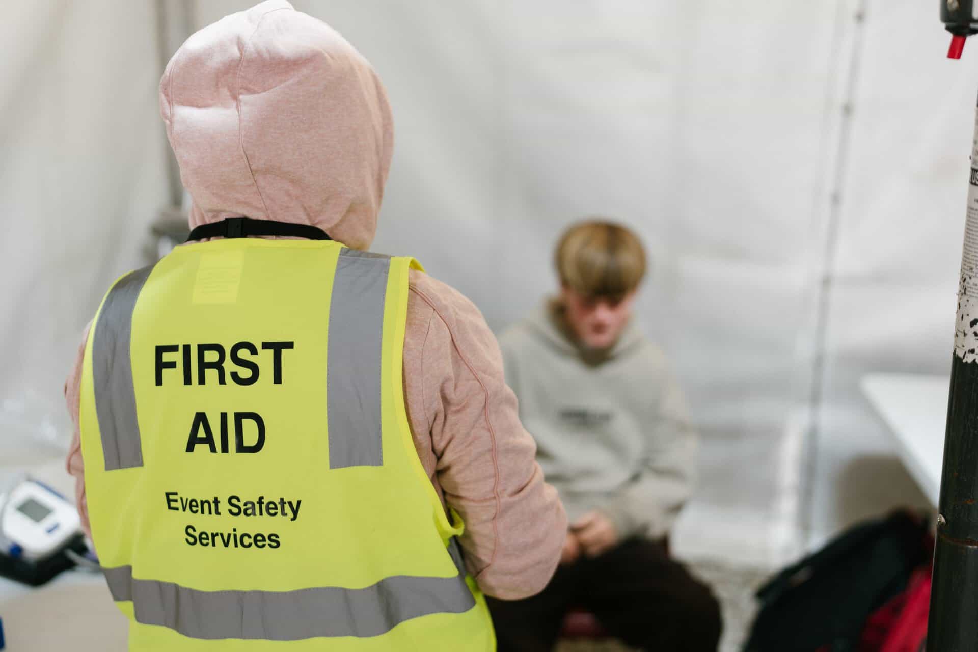 An emergency responder wearing a high-visibility vest with "FIRST AID" and "Event Safety Services" printed on the back, attending to a young person seated inside a safety tent.