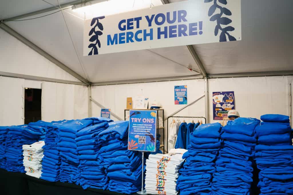 Merchandise booth at KYCK 2026 event with stacks of blue T-shirts and signage for merchandise sales.
