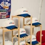 Collection of KYCK 2026 branded caps with white and navy blue design, displayed on wooden shelves at the event.