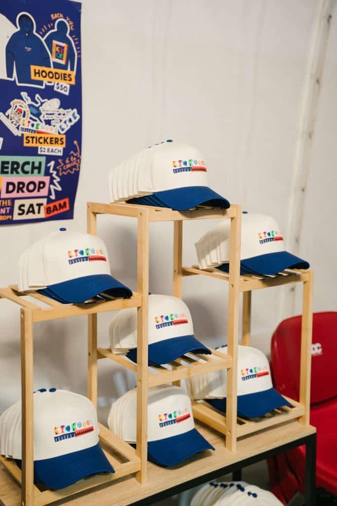 Collection of KYCK 2026 branded caps with white and navy blue design, displayed on wooden shelves at the event.