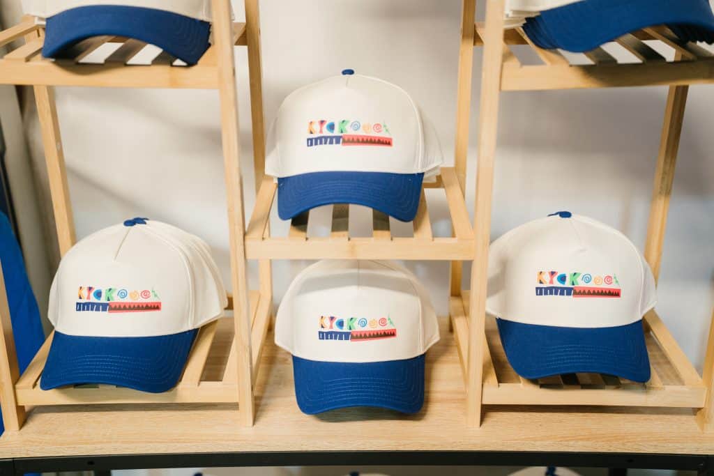 White and blue KYCK 2026 caps displayed on wooden shelves, featuring colourful KYCK logo, for youth church event in Australia.
