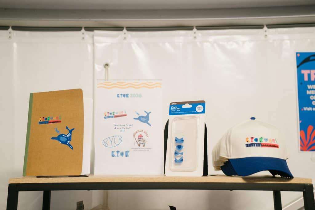 Display of KYCK 2026 event merchandise including notebooks, caps, and promotional materials on a table.