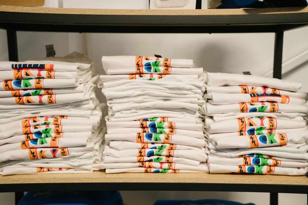 Stacked white KYCK 2026 event T-shirts with colourful logo designs on a wooden shelf.